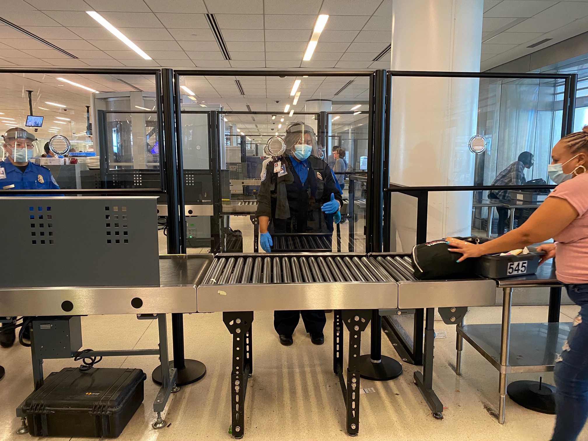 TSA officers in masks, gloves and face shields standing behind an acrylic barrier as travelers divest their items into a checkpoint bin for X-ray screening. (TSA photo)